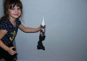 Uśmiechnięta dziewczynka trzyma srebrną rakietę pokazując ją, jakby startowała w górę